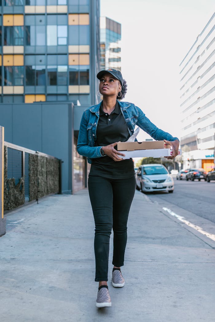 Delivery woman wearing a denim jacket walking on a city sidewalk carrying pizza boxes. Urban job scene.