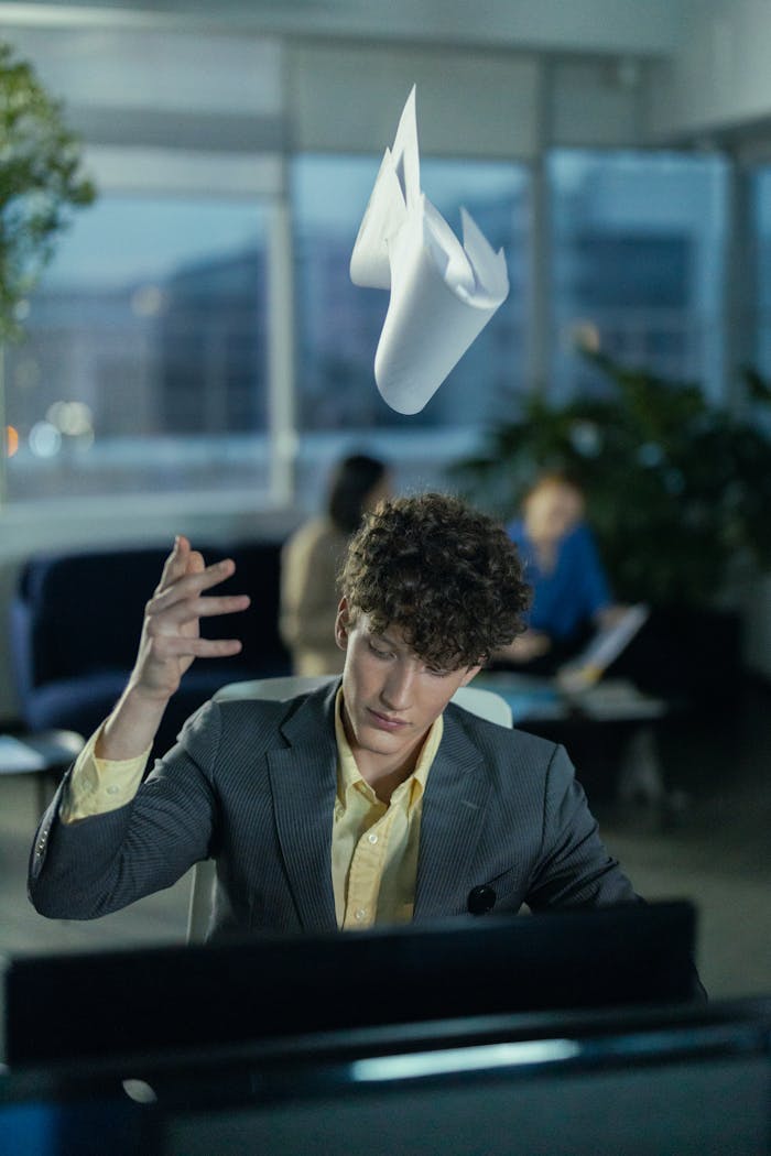 Young office worker in gray suit frustrated, tossing papers indoors.